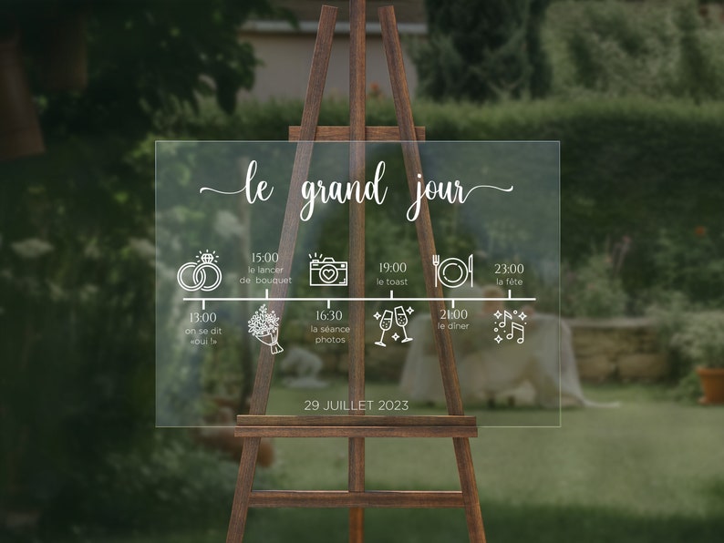 May include: A clear acrylic sign with a white outline of a wedding timeline. The sign includes the text "le grand jour" and lists the times for various wedding events, including the ceremony, photos, dinner, and dancing. The date "29 JUILLET 2023" is printed at the bottom of the sign.