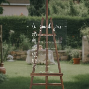 May include: A clear acrylic sign with a white calligraphy script that reads "le grand jour" and a wedding day schedule. The sign is on an easel and the background is a blurred image of a garden.