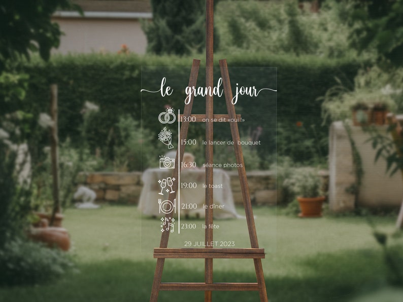 May include: A clear acrylic sign with a white calligraphy script that reads "le grand jour" and a wedding day schedule. The sign is on an easel and the background is a blurred image of a garden.