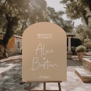 May include: A tan arch-shaped welcome sign with white text that reads "Bienvenue au mariage de Alice + Bastien 27.08.23". The sign is on a wooden easel in front of a stone building and lush greenery.