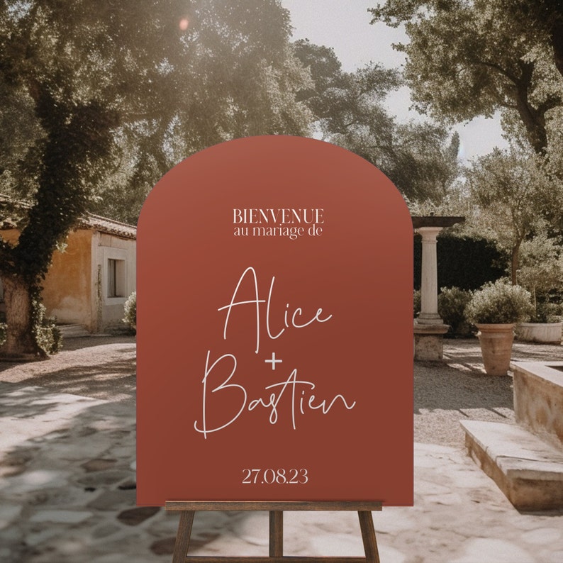 May include: A terracotta-colored archway sign with white text announcing the wedding of Alice and Bastien on August 27, 2023. The sign reads "Bienvenue au mariage de Alice + Bastien 27.08.23".