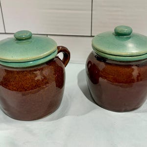 May include: Two matching ceramic jars with rounded bodies and dark brown glossy glaze. Each jar features a single side handle and a removable lid with a smooth, light green finish. The containers have a wide base and a narrow opening.