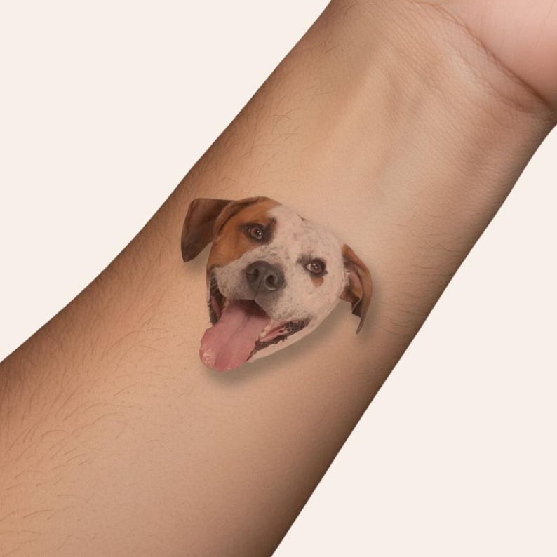 May include: A temporary tattoo of a smiling dog's face, featuring brown and white markings, with its tongue sticking out. The tattoo is applied to the arm, showcasing the design's details and colors.