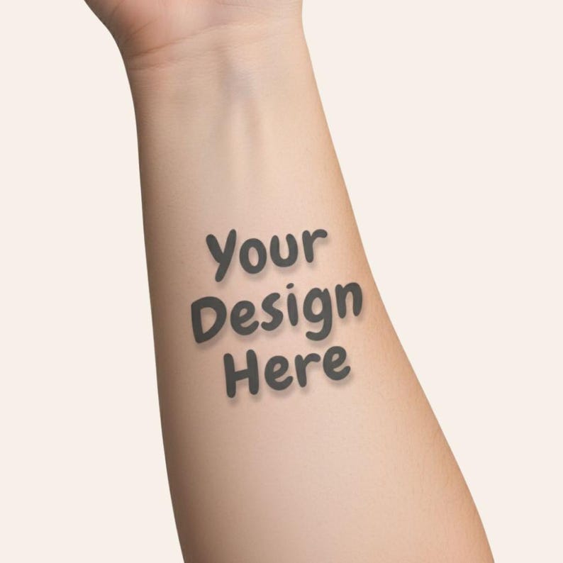 May include: A temporary tattoo on a forearm with the text "Your Design Here" in a dark gray, slightly shadowed font. The skin tone is light, and the background is a solid, neutral color.