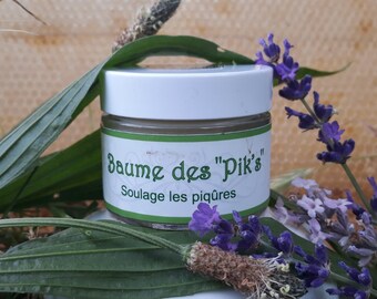 May include: A small white jar with a green label that reads "Baume des "Pik's" Soulage les piqûres". The jar is surrounded by green leaves and purple lavender flowers.