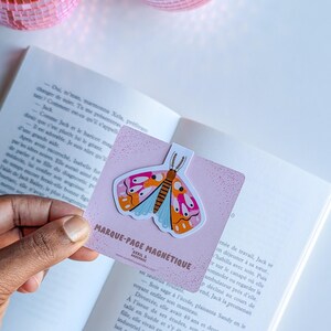 Peut inclure: Un marque-page magnétique rose avec une illustration de papillon coloré. Le papillon a des ailes orange, roses et bleues. Le marque-page est fixé aux pages d'un livre. Le texte sur le marque-page est "MARQUE-PAGE MAGNETIQUE".