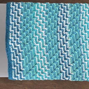 Op de afbeelding: Een gehaakt deken met een chevron-patroon in tinten blauw en wit.