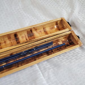 Puede incluir: Caja de madera rectangular abierta, que revela varias cañas de pescar azules. La caja, con tapa con bisagras, es de madera marrón claro con un patrón de vetas natural. Las cañas de pescar son de color azul oscuro. Fondo blanco.