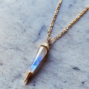 Könnte beinhalten: Eine goldfarbene Halskette mit einem Anhänger in Form eines Kristallpunkts. Der Kristall hat einen Farbverlauf von klar zu blau. Die Halskette und die Kappen des Anhängers sind ebenfalls goldfarben. Der Anhänger ist etwa 3 cm lang.