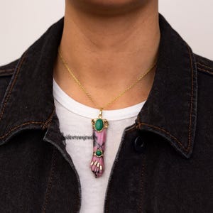 Könnte beinhalten: Eine goldfarbene Halskette mit einem Anhänger in Form einer Hand, gefertigt aus rosa und schwarzem Stein. Der Anhänger ist mit zwei grünen Edelsteinen verziert und hängt an einer feinen Goldkette. Die Halskette wird über einem weißen Oberteil und einer schwarzen Jeansjacke getragen.