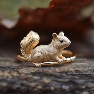 May include: A gold-colored ring featuring a detailed squirrel design. The squirrel has a bushy tail and small, dark eyes. The ring is set on a wooden surface, showcasing the intricate details of the jewelry.