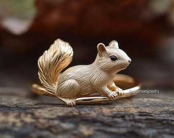 May include: A gold-colored ring featuring a detailed squirrel design. The squirrel has a bushy tail and small, dark eyes. The ring is set on a wooden surface, showcasing the intricate details of the jewelry.