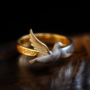 Könnte beinhalten: Ein silberner und goldener Ring mit einem Vogeldesign. Der Vogel hat silberne Federn und goldene Flügel, Schnabel und Füße. Das Ringband ist goldfarben mit einem strukturierten Muster auf der Außenseite und einem silbernen Innenband. Dunkler Hintergrund, Holzoberfläche.