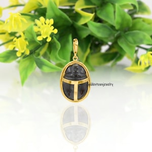 Puede incluir: Colgante dorado con un centro de piedra ovalada gris oscuro, con un diseño de escarabajo. El colgante tiene un marco dorado y un diseño de cruz dorada. El fondo incluye hojas verdes y flores amarillas.