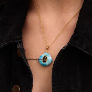 May include: A gold chain necklace with a pendant featuring a light blue stone with a dark brown center. The pendant has a unique, irregular shape. The necklace is worn against a dark background.