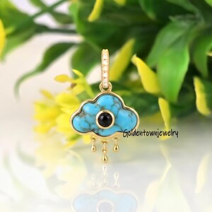 Può includere: Un ciondolo in oro a forma di nuvola con un inserto in turchese e una pietra circolare nera centrale. Il ciondolo presenta tre piccole gocce d'oro pendenti sul fondo e un piccolo gancio sulla parte superiore. Il testo Goldtownjewelry è visibile.