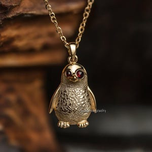 Op de afbeelding: Een goudkleurige pinguïnhangerketting. De pinguïn heeft rode ogen en gedetailleerde details op zijn lichaam. De ketting is ook goudkleurig. De hanger hangt aan een delicate ketting.