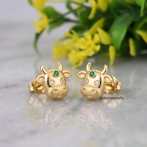Könnte beinhalten: Ein Paar goldene Ohrstecker in Form von Kuhköpfen. Jeder Ohrstecker ist mit einem kleinen grünen Edelstein auf der Stirn, zwei klaren weißen Steinen als Augen und einer texturierten, matten Oberfläche im Schnauzenbereich versehen.