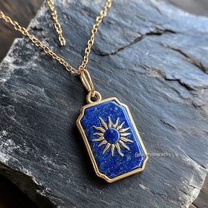 Puede incluir: Un collar de cadena dorada con un colgante rectangular. El colgante presenta una piedra azul oscuro con un diseño de sol dorado. El colgante está enmarcado en oro y descansa sobre una superficie gris oscuro.