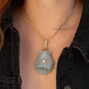 Puede incluir: Collar de cadena dorada con un colgante de concha marina azul claro. El colgante tiene un diamante central y detalles dorados. El collar se lleva sobre una chaqueta vaquera negra.