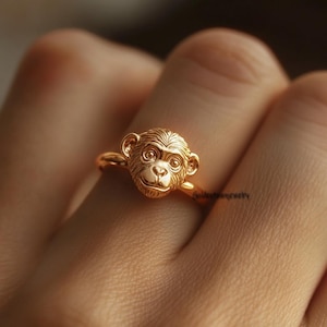Peut inclure: Une bague dorée représentant un visage de singe détaillé. La bague est portée au doigt, mettant en valeur le design complexe des traits du singe. La bague est un bijou.