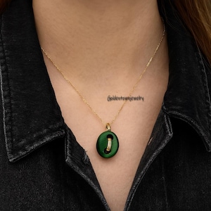 May include: A gold-colored chain necklace with a dark green pendant. The pendant is a button shape with a gold-colored ring in the center. The necklace is worn against a black background.
