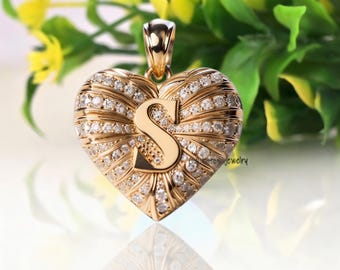 Puede incluir: Un colgante en forma de corazón de oro con una letra S central en acabado pulido. La superficie está decorada con líneas radiantes de pequeñas piedras redondas transparentes engastadas en el metal dorado. Un asa de oro sujeta la parte superior.