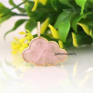 May include: A cloud-shaped pendant featuring a smooth, pale pink rose quartz stone set in a gold-toned metal frame. The top bail is accented with small, clear, sparkling stones. The text Goldentownjewelry is printed on the right side.