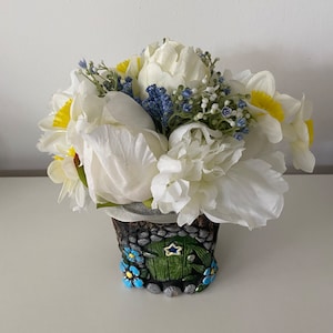 May include: A white and yellow flower arrangement in a small, ceramic, cottage-style planter. The planter is decorated with a green door and blue flowers.