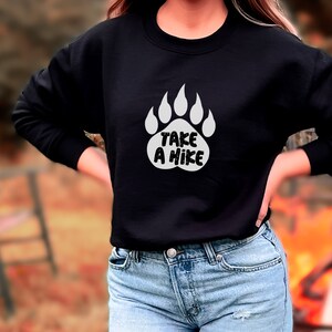 Pode incluir: Camisola de moletom preta com uma pegada de urso branca e o texto "Take a Hike" impresso na frente.