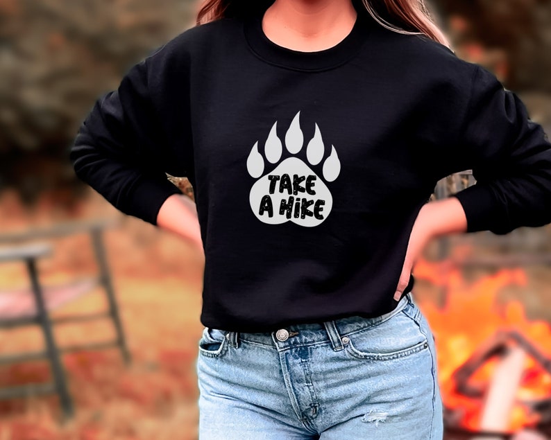 Pode incluir: Camisola de moletom preta com uma pegada de urso branca e o texto "Take a Hike" impresso na frente.