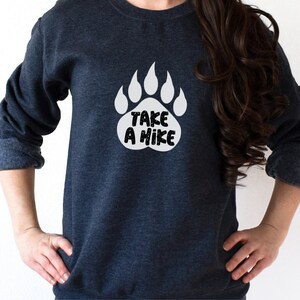 Pode incluir: Uma camisa de moletom azul escura com um gr&aacute;fico de impress&atilde;o de pata de urso branca que diz "Take a Hike".