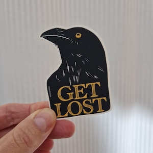 Può includere: Adesivo nero e bianco di un corvo con occhi gialli e il testo "GET LOST" in lettere dorate.