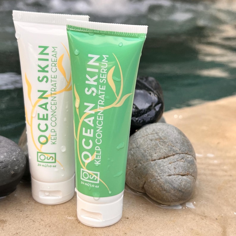 Może przedstawiać: Dwa białe tuby z kremem i serum koncentratem z alg morskich Ocean Skin. Tuby mają pojemność 30 ml/1 oz i są w zielono-białym kolorze. Tuby leżą na stercie szarych kamieni.