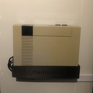 May include: A beige and black Nintendo Entertainment System (NES) console mounted on a wall with a black bracket. The bracket has the Nintendo logo printed on it.