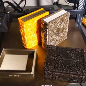May include: Three 3D printed book-shaped boxes. The boxes are in gold, silver, and black. The gold box has the word "SPIRE" printed on the front. The boxes are sitting on a brown surface.