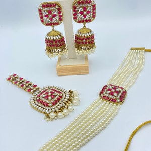 Puede incluir: Un conjunto de joyas de novia indias en rosa y dorado, con un collar de gargantilla de perlas, un tikka a juego y un par de pendientes. Los pendientes tienen un diseño cuadrado con piedras rosas y una campana colgante.