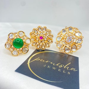 Puede incluir: Tres anillos de oro con piedras blancas y una piedra central. Las piedras centrales son verde, rosa y blanca. Los anillos están sobre una superficie blanca con una tarjeta negra que dice "punisha jewels".