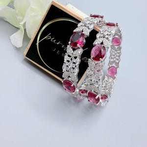 May include: Two silver bangles, each set with oval and round red gemstones and small, clear stones. The jewellery is displayed on a black and gold box with the text "pure jewels".