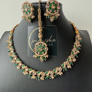 May include: A gold-toned jewelry set featuring a necklace, earrings, and a headpiece. The set is adorned with green and clear gemstones. The necklace and earrings have a floral design. The headpiece has a central pendant with a matching design. The jewelry is displayed on a black mannequin.