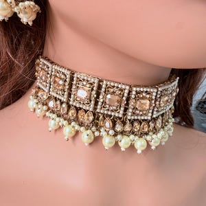 Könnte beinhalten: Ein goldfarbenes Choker-Halsband mit quadratischen und tropfenförmigen bernsteinfarbenen Edelsteinen und kleinen weißen Perlen. Die Halskette ist mit kleinen, klaren Kristallen verziert.