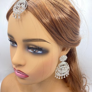 May include: A silver and white bridal headpiece set with a large, round, jeweled pendant and matching earrings. The pendant and earrings feature a circular design with multiple layers of sparkling stones.