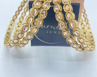 Op de afbeelding: Set van vier gouden armbanden met heldere ovale strasssteentjes. De armbanden zijn op elkaar gestapeld en liggen op een zwarte doos met het zichtbare woord "JEWE".