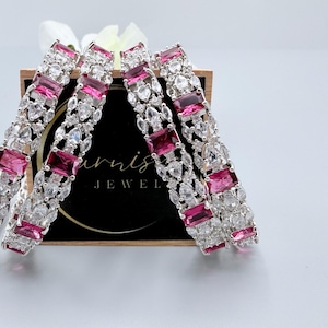 May include: Four silver bangles with alternating pink and clear gemstones. The rectangular pink stones are interspersed with clear, teardrop-shaped stones. The bangles are displayed in front of a black background with the text "Furnish Jewel".