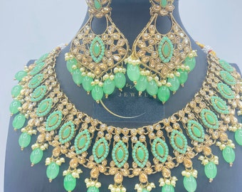 May include: A gold necklace and earrings set with green and clear stones and pearls. The necklace has a layered design with a central row of large green stones and a dangling row of smaller green stones. The earrings are teardrop shaped with a similar design.