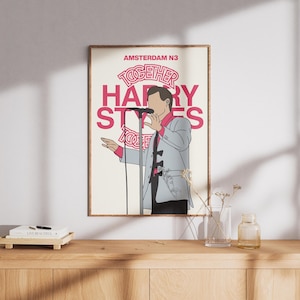 Op de afbeelding: Een ingelijste poster met een illustratie van een zanger op het podium. De tekst "AMSTERDAM N3 TOGETHER HARRY STYLES" staat in roze en wit. De zanger draagt een lichtblauw pak en houdt een microfoon vast.