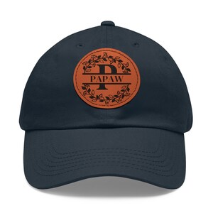 May include: A navy blue baseball cap with a brown leather patch featuring a floral wreath and the letter "P" inside, with the word "PAPAW" below it.