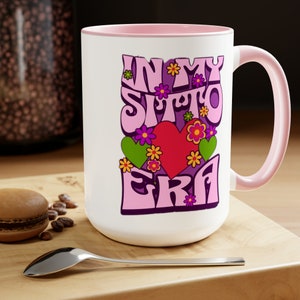 May include: White ceramic mug with a pink rim and handle. The mug features a purple and pink graphic with the text "In My Sitto Era" and a red heart with green and pink flowers.