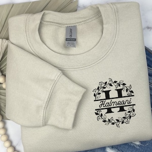May include: A light beige sweatshirt with a black floral monogram design featuring the letter "H" and the name "Halmeoni".