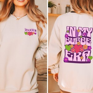 May include: A cream-colored sweatshirt with a pink and purple graphic on the front that says "BUBBE" and a pink and purple graphic on the back that says "IN MY BUBBE ERA".
